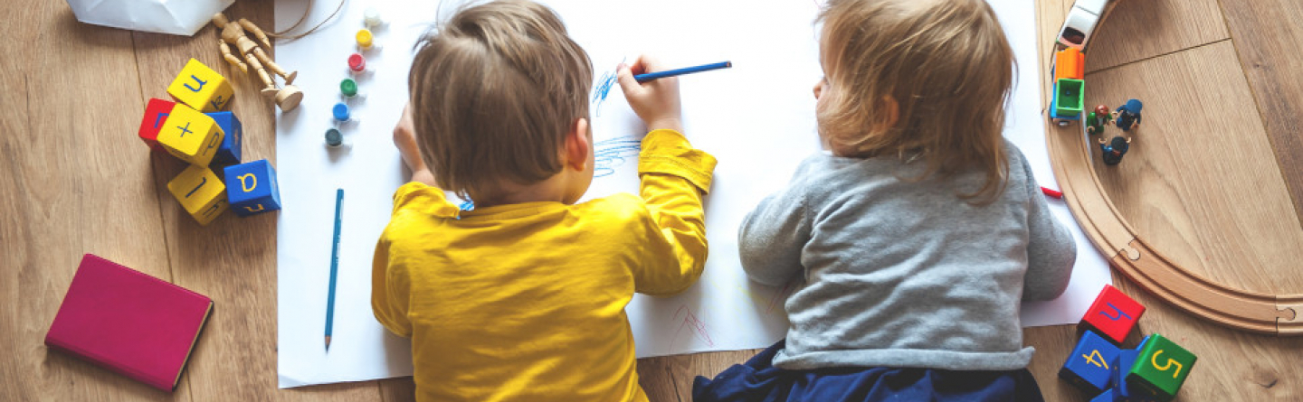 Zwei Kleinkinder liegen am Boden und Zeichnen etwas auf einen Malblock. 