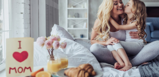 Im Hintergrund ist eine Mutter zu sehen, welche ihr Kind umarmt und im Vordergrund ist ein Frühstück zu sehen, welches eine Karte mit den Worten "I love mom" enthält. 